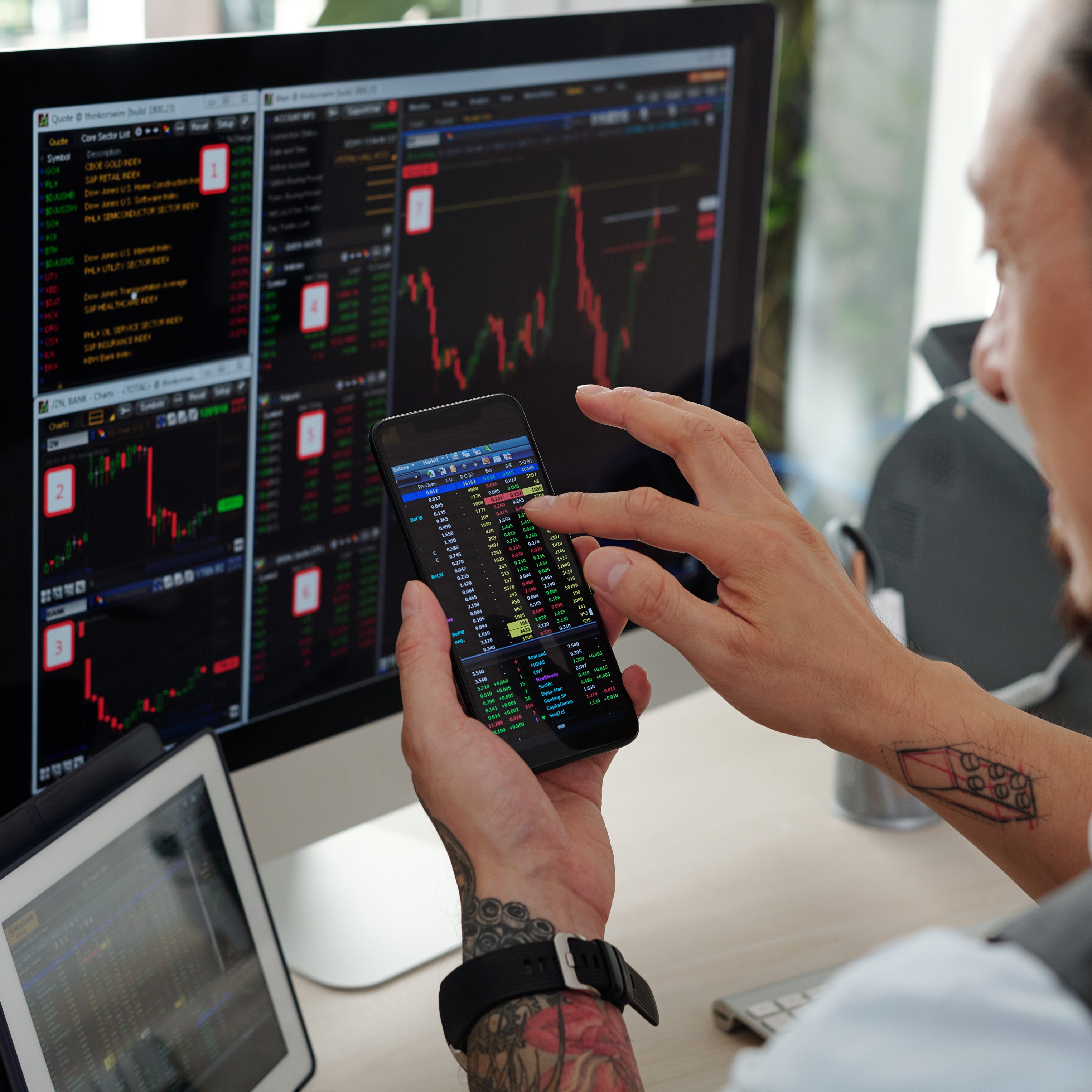 Trader checking stock market charts on computer screens and buying stocks via application on smartphone